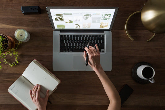laptop on desk with a person hovering hand over keyboard. An open notepad, cup of coffee, plant, lamp and candle also on desk.