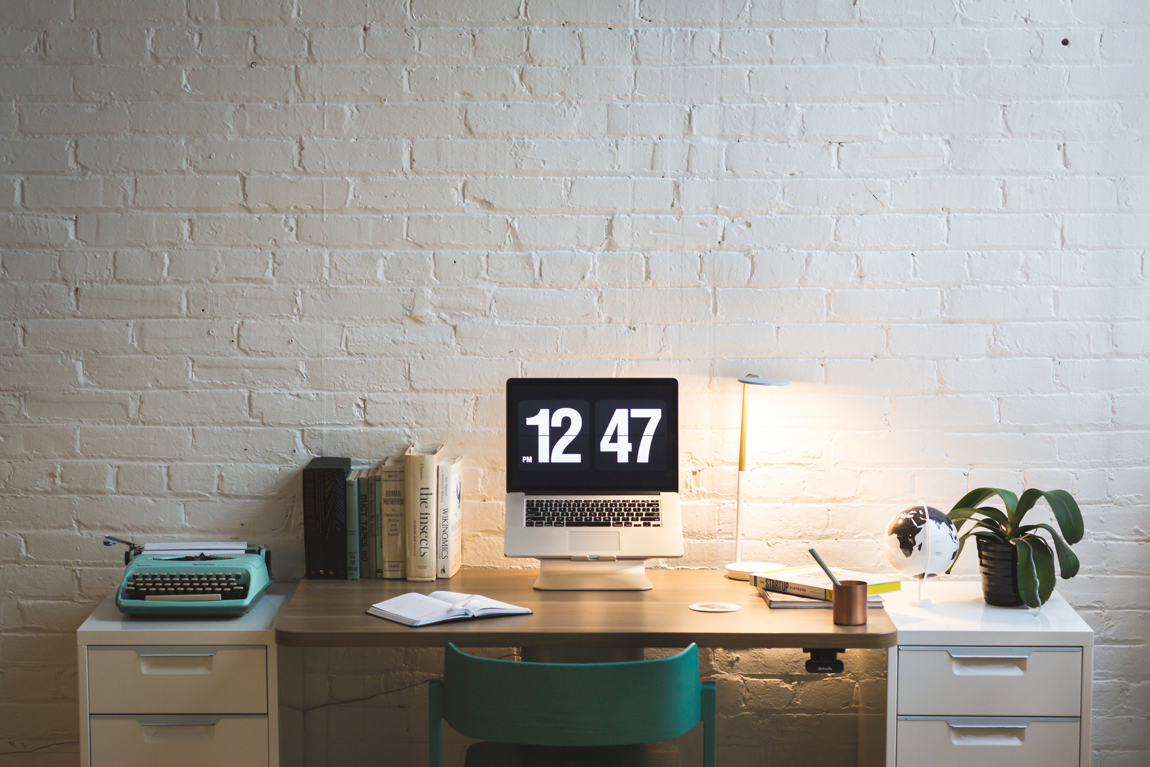 desk with computer, lamp, plant and books, an open notepad and teal chair against a white brick wall