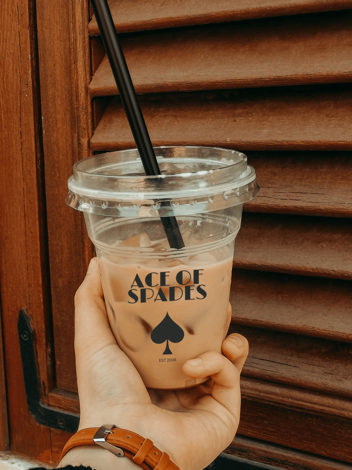 Hand holding a clear cup with a black straw labeled 'Ace of Spades' against a wooden background